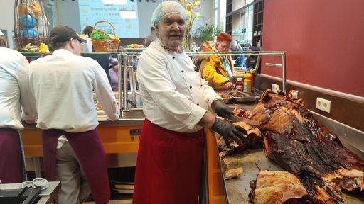 Ein Mann in Koch-Kleidung steht hinter einer Essensausgabe neben einem Spanferkel.