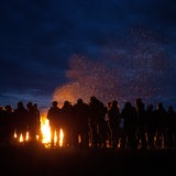 Eine Gruppe Menschen steht am Abend vor einem großen Lagerfeuer