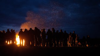 Eine Gruppe Menschen steht am Abend vor einem großen Lagerfeuer