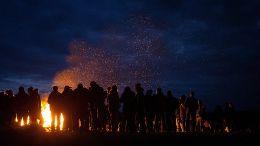 Eine Gruppe Menschen steht am Abend vor einem großen Lagerfeuer