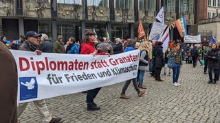 Menschen halten ein Banner auf dem Marktplatz auf dem "Diplomaten statt Granaten" steht.