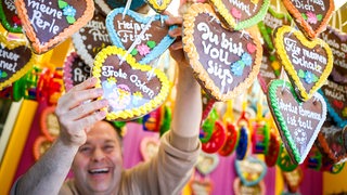 Lebkuchenherzen mit netten Botschaften hängen im Vordergrund. Im Hintergrund steht ein Mann und hält eines der Herzen in die Kamera.