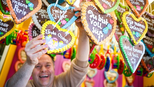 Lebkuchenherzen mit netten Botschaften hängen im Vordergrund. Im Hintergrund steht ein Mann und hält eines der Herzen in die Kamera.