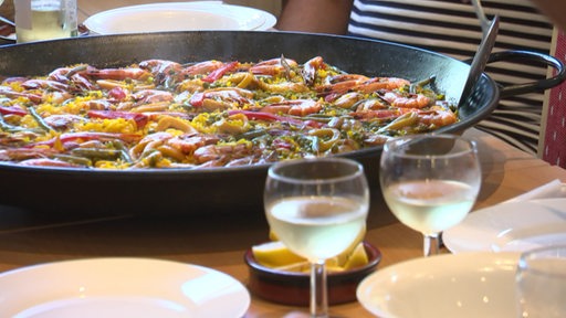 Eine Pfanne mit zubereiteter Paella steht auf einem Tisch mit Weingläsern im Vordergrund.