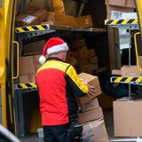 Ein DHL-Mitarbeiter mit einer Weihnachts-Mütze auf dem Kopf holt Pakete aus seinem Wagen.