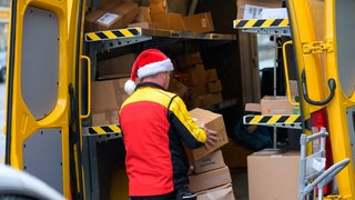 Ein DHL-Mitarbeiter mit einer Weihnachts-Mütze auf dem Kopf holt Pakete aus seinem Wagen.