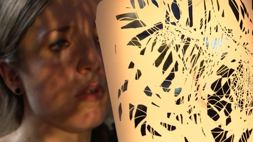 Eine Künstlerin hält ihre Kunst vor ihr Gesicht. Licht scheint durch das zerschnittene papier.