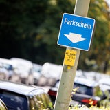 Ein Schild weist am Straßenrand neben geparkten Fahrzeugen auf die Position eines Parkscheinautomaten hin. 
