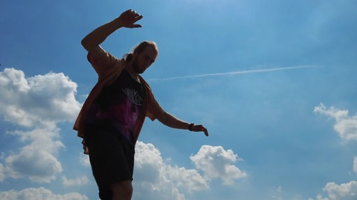 Es ist die Silhouette einer Person vor einem blauen Himmel zu sehen. Sie balanciert mit ausgestreckten Armen auf etwas. 