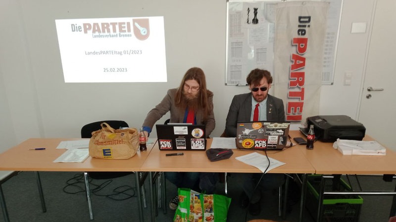 Zwei Männer hinter zusammengerückten Tischen vor dem Logo der Partei "Die Partei"