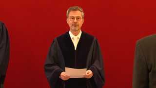 Ein Richter des Staatsgerichtshofs steht vor einer roten Wand und verliest ein Urteil.