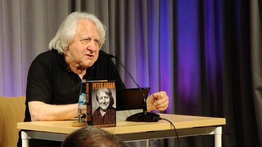 Ein Mann mit grauen Haaren sitzt an einem Tisch auf einer Bühne, auf dem Tisch stehen ein Mikorofon und ein Busch mit der Aufschrift Peter Urban.