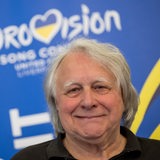 Ein Mann, etwas älter, lächelt in die Kamera. Im Hintergrund ein Plakat mit der Aufschrift Eurovisiion Song Contest.