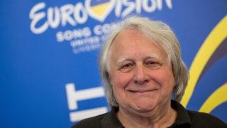 Ein Mann, etwas älter, lächelt in die Kamera. Im Hintergrund ein Plakat mit der Aufschrift Eurovisiion Song Contest.