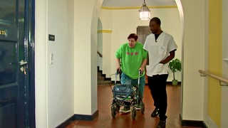 Ein Pfleger läuft zusammen mit einer Seniorin über einen Flur