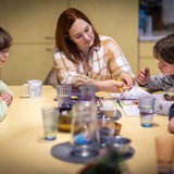 Eine Frau sitzt mit Kindern an einem Tisch in einer Küche und hilft bei Hausaufgaben und plant den Familienalltag.