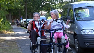 Zwei Seniorinnen sitzen nebeneinander auf einem Fahrrad und treten in die Pedale.