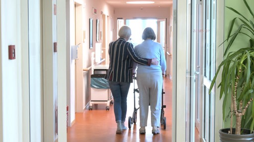 Eine Pflegerin stützt eine ältere Dame in dem Flur einer Pflegeeinrichtung.