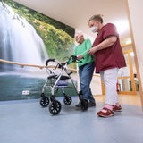 Eine alte Frau mit einem Rollator und einer Frau in Pflegekraft-Kleidung läuft einen Flur entlang.