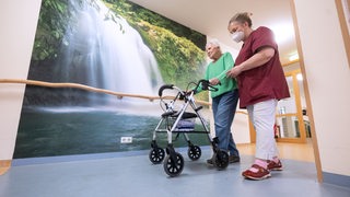 Eine alte Frau mit einem Rollator und einer Frau in Pflegekraft-Kleidung läuft einen Flur entlang.