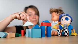 Bunte Holzbausteine werden von einer Frau gestapelt, im Hintergrund sind zwei Kuscheltiere
