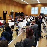 Ein Dirigent steht mit erhobenen Armen vor Musikern, die an Notenständern sitzen.