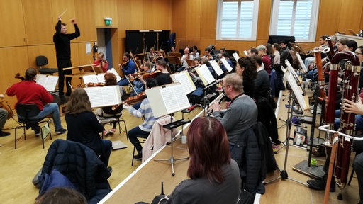 Ein Dirigent steht mit erhobenen Armen vor Musikern, die an Notenständern sitzen.