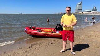 Ein Mann steht am Strand vor einem Schlauchboot und einer Skyline.