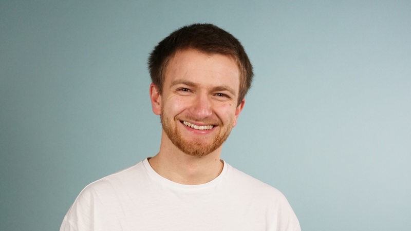 Ein junger Mann steht vor einer blauen Wand und lächelt in die Kamera.