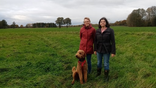 Zwei junge Frauen und ein Hund stehen auf einem weiten Feld.