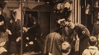 Männer in Uniformen mit Hakenkreuz-Aufnähern geben Sachen raus durch ein kaputtes Schaufenster