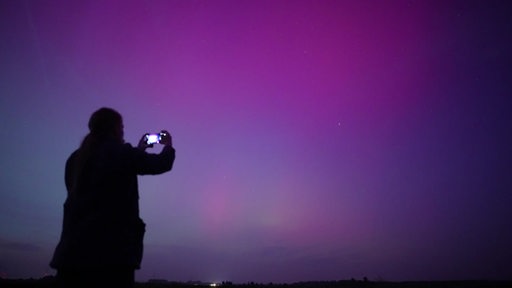 Es ist eine dunkle Silhouette einer Person, welche den rosa-lilanen Himmel fotografiert, zu sehen.