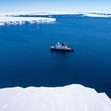 Ein Schiff fährt über blaues, ruhiges Wasser. Im Hinter- und Vordergrund sind große Eisschollen zu sehen.