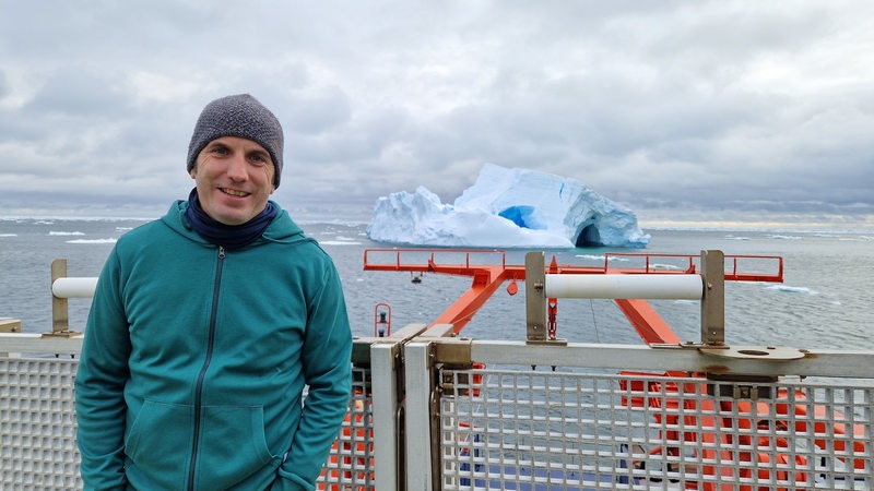 Klimaforscher Marcus Gutjahr posiert vor einem Eisberg in der Antarktis