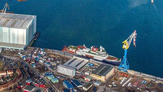 Eine Illustration zeigt aus der Luft ein Schiff am Kai in einem Hafengebiet.