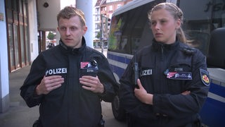 Die zwei Polizeikommissare Lars Zschernig und Anna Meyer stehen vor einem Polizeiauto. 