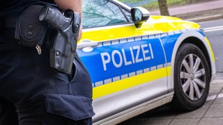 Ein Polizist mit einer Waffe am Gürtel steht vor einem Polizeiauto.
