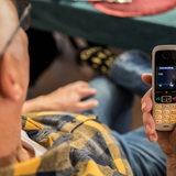 Ein älterer Menschen sitzt in seiner Wohnung und hält ein Mobiltelefon in der Hand. Auf dem Display des Telefons ist die 110 zu lesen.