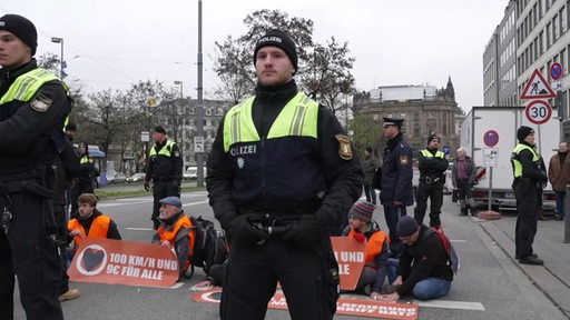 Zu sehen ist ein Polizist, welcher vor einer Reihe von Demonstranten der letzten Generation steht.