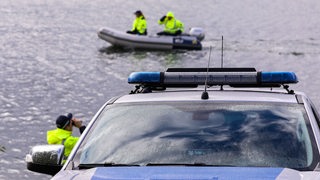 Ein Polizeiauto am Ufer vor einem Fluss, im Hintergrund Polizisten mit Westen in Schlauchbooten.