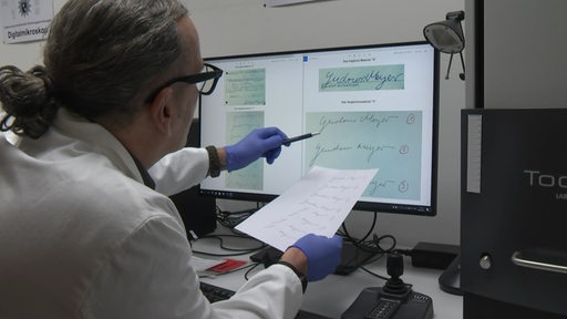 Ein Handschriftenprüfer hält ein Blatt Papier mit einer Unterschrift vor einen Computer-Monitor und vergleicht die Schriften.