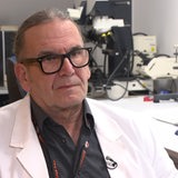 Ein Mann mit langen grauen Haaren, die er zu einem Zopf gebunden hat und einer großen schwarzen Brille schaut in die Kamera. Im Hintergrund sind Messapparate zu sehen