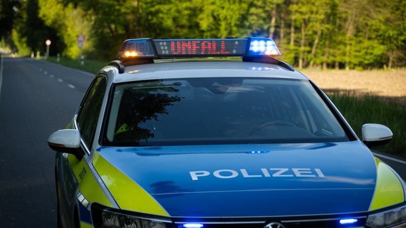 Ein Polizeiauto steht auf einer Landstraße. Auf einer Leuchttafel auf dem Dach des Autos steht "Unfall".