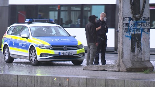 Ein Streifenwagen steht auf Vorplatz vom Bremer Hauptbahnhof. Davor steht ein Polizist bei einer augenscheinlichen Kontrolle von einer Person. Im Hintergrund fährt eine Straßenbahn.