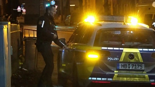 Ein Polizist steht neben einem Streifenwagen. Die Blaulichter leuchten im Dunkeln.