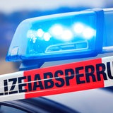 Ein Absperrband der Polizei mit der Aufschrift "Polizeiabsperrung". Im Hintergrund ist das Blaulicht eines Polizeiautos zu sehen.