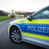 Ein Polizeiwagen steht auf einer Landstraße. Im Hintergrund ist ein Rettungshubschrauber zu sehen.