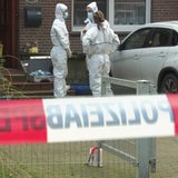 Hinter einer Polizeiabsperrung stehen drei Beamte der Polizei in weißer Kleidung vor einem haus 