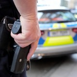 Ein Polizist steht mit seiner Hand auf der Schusswaffe vor einem Einsatzfahrzeug der Polizei.