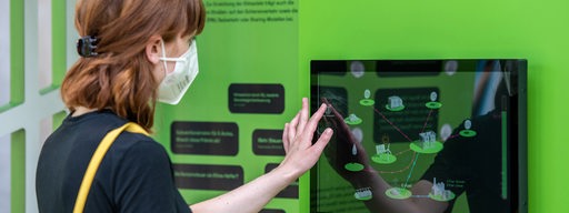 Eine junge Frau mit Maske tippt auf einen Touchscreen an einer grünen Wand.